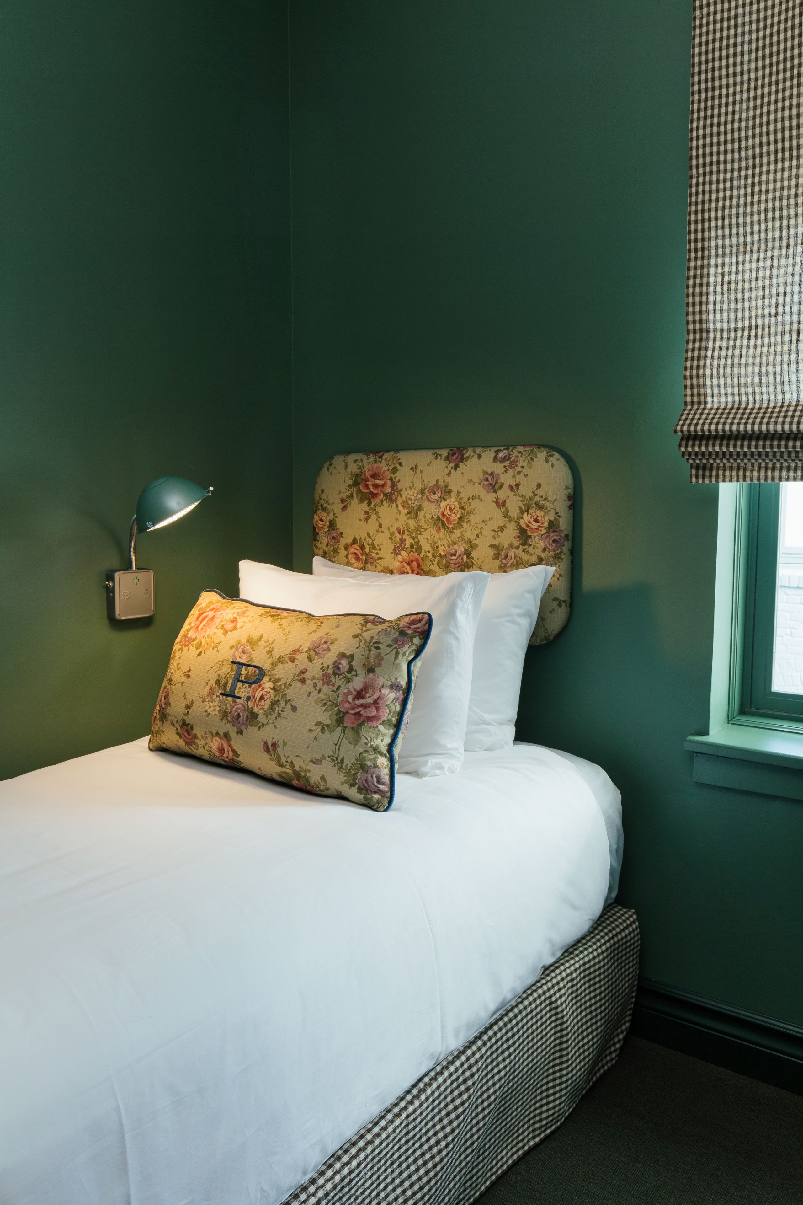 A neatly made bed with floral headboard and pillows, a bedside lamp, and a window with a checkered blind.