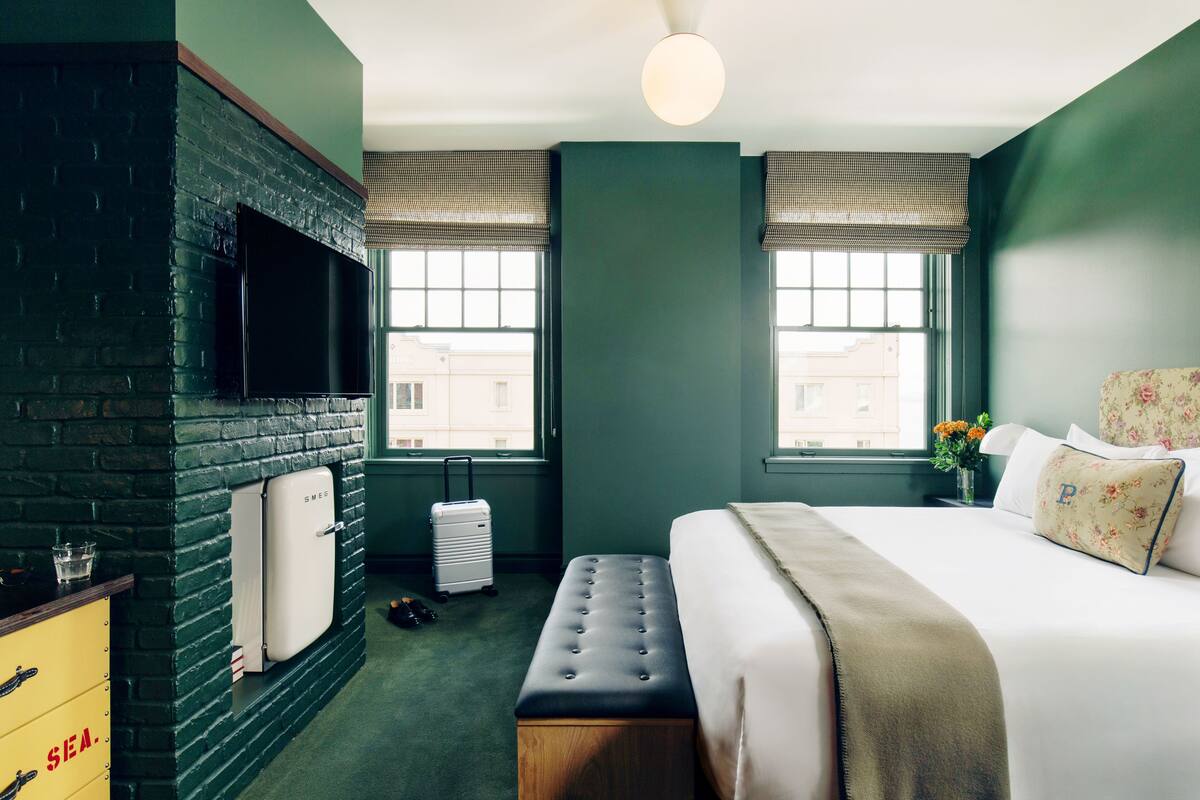 A hotel room with a brick wall, a flat-screen TV, a bed with floral pillows, and a bench.