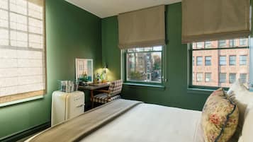 A room with a bed, a desk with a chair, a vintage refrigerator, and a window with a view of buildings.