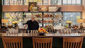 Un espace bar avec un barman, une grande fenêtre donnant sur la ville et des étagères remplies de bouteilles et de verres.