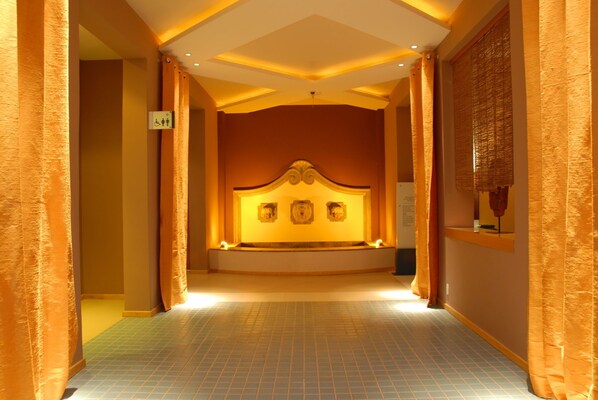 A corridor with a central seating area featuring a headboard with three decorative panels, leading to a room with a tiled floor and warm lighting. - Blue Tree Towers Sao Luis (São Luís)