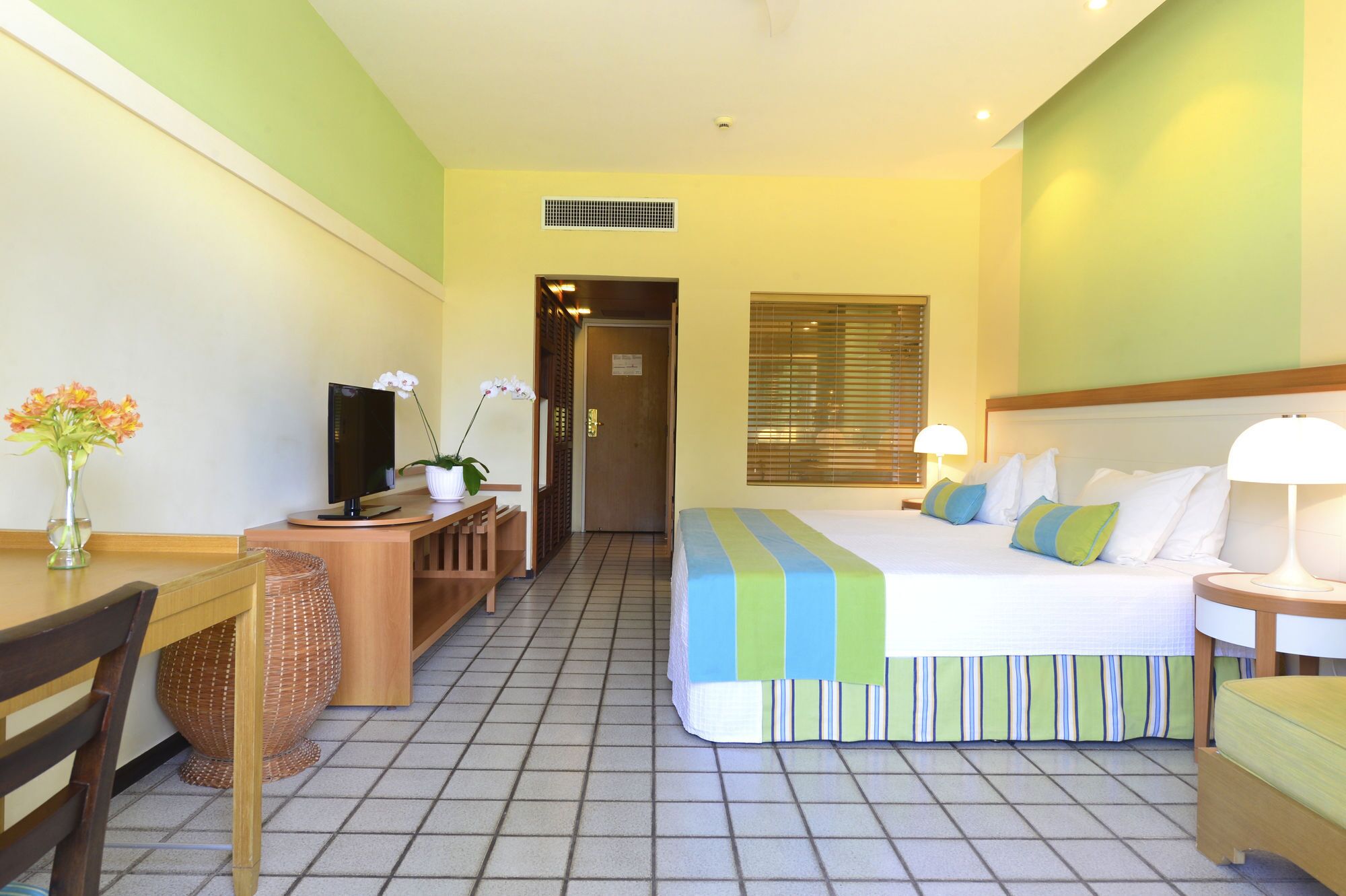 Una habitación de hotel con cama, escritorio, silla, televisión y ventana con persianas.