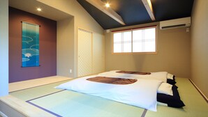 Ein Zimmer mit Tatami-Matten, zwei Betten mit weißer und brauner Bettwäsche, einem Fenster mit Jalousien, einer Klimaanlage und einer Wand mit einer dekorativen Wandhängung.