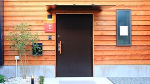 Un bâtiment en bois avec une porte noire, une enseigne indiquant « COTO Fuzhimari 3 », et une petite plante placée à côté de l’entrée.