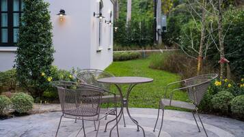 Un patio con una mesa redonda y dos sillas, rodeado de follaje y un edificio blanco con ventanas y luces en la pared.