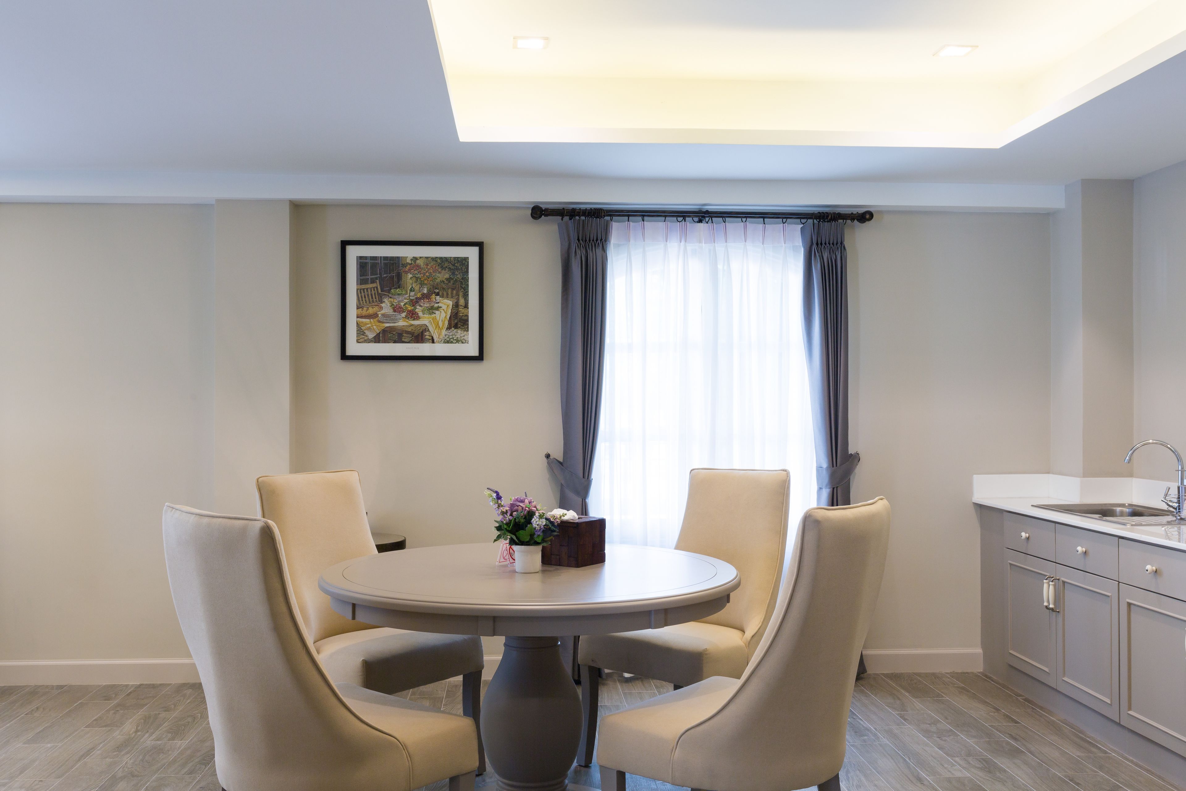 A dining area with a round table and four chairs, a small vase with flowers, a framed picture on the wall, and a window with curtains.