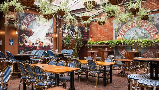 Un comedor interior con sillas de mimbre y mesas de madera, plantas colgantes y un mural en la pared.