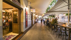 Un café al aire libre con mesas y sillas, una zona de bar con botellas y un cartel que dice 'Mussels Stella'.