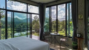 A bedroom with a large bed, a small table, two chairs, and a view of a mountainous landscape through large windows. - Phu Pha View Doi (Samoeng)