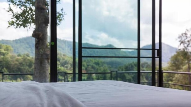 A bed with a white sheet in a room with large windows, offering a view of a forested mountain range. - Phu Pha View Doi (Samoeng)