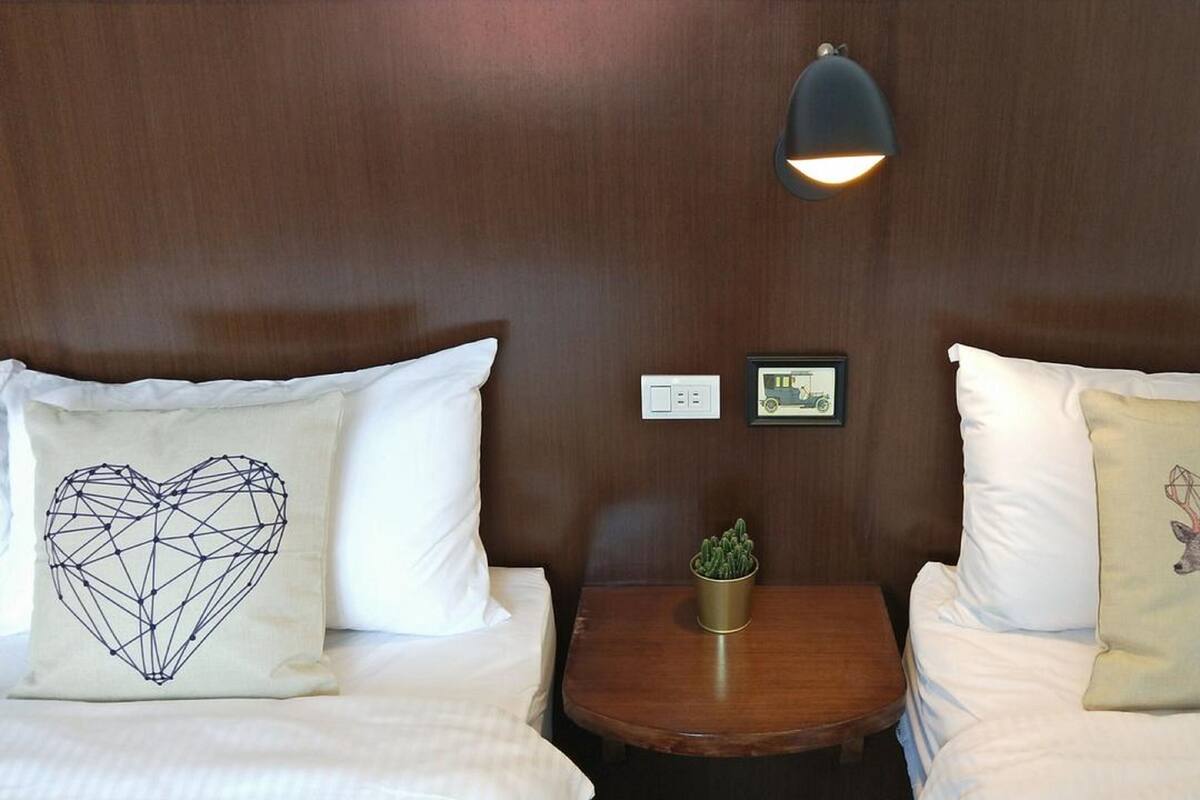 A hotel room with two beds, each with a heart-shaped and a deer-themed pillow, a small table with a plant, and a wall-mounted light fixture.