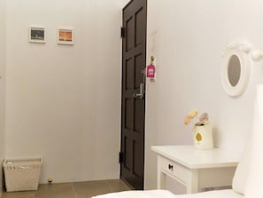A room with a white dresser, a mirror, and a small vase with flowers. There is a door with a black frame and a small basket on the floor.