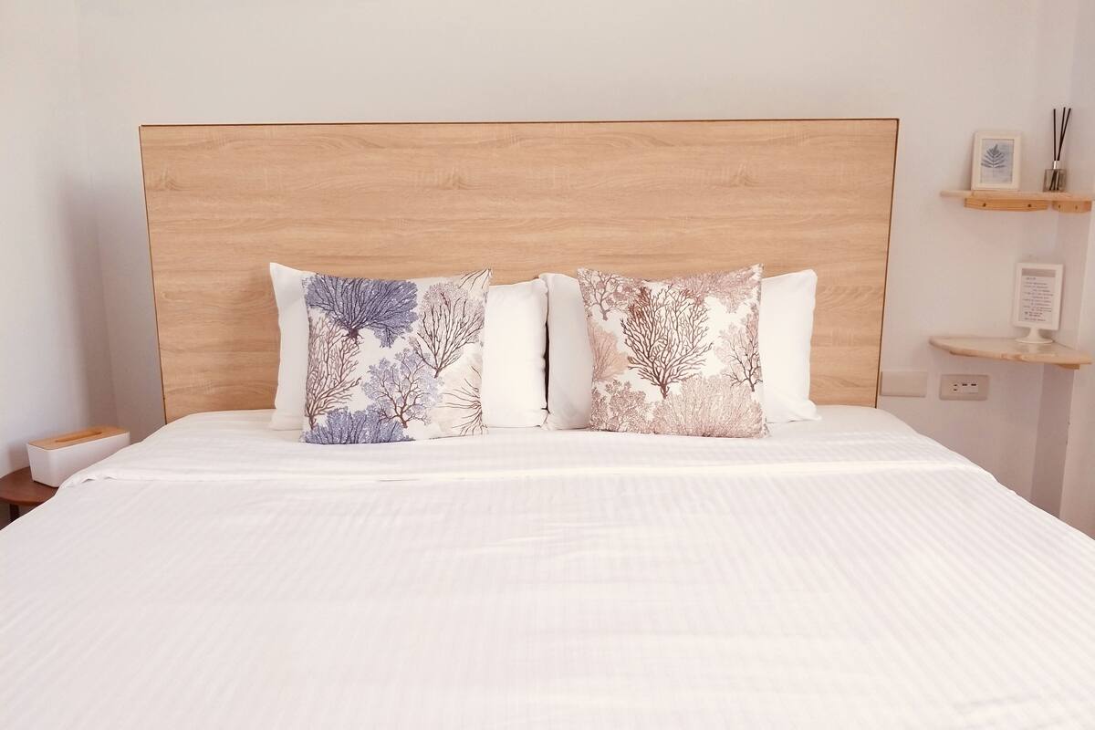 A neatly made bed with two decorative pillows, a wooden headboard, a bedside table with a tissue box, and a small shelf with a framed picture and a candle.