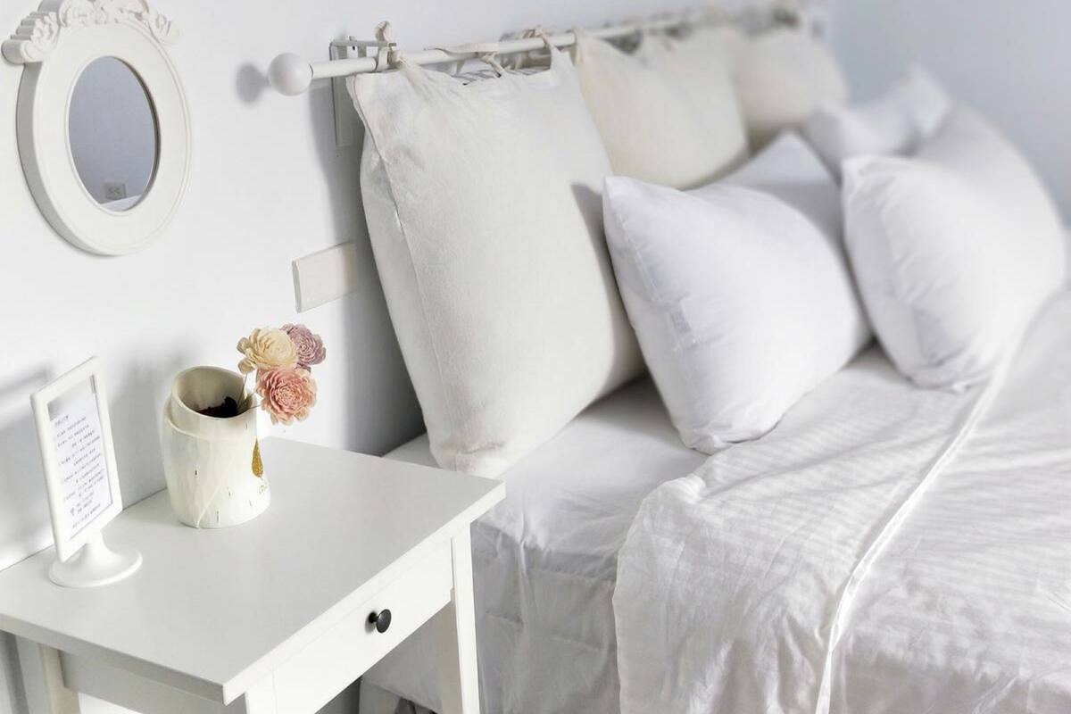 A white bedroom with a bed, a nightstand, a mirror, and a vase with flowers.