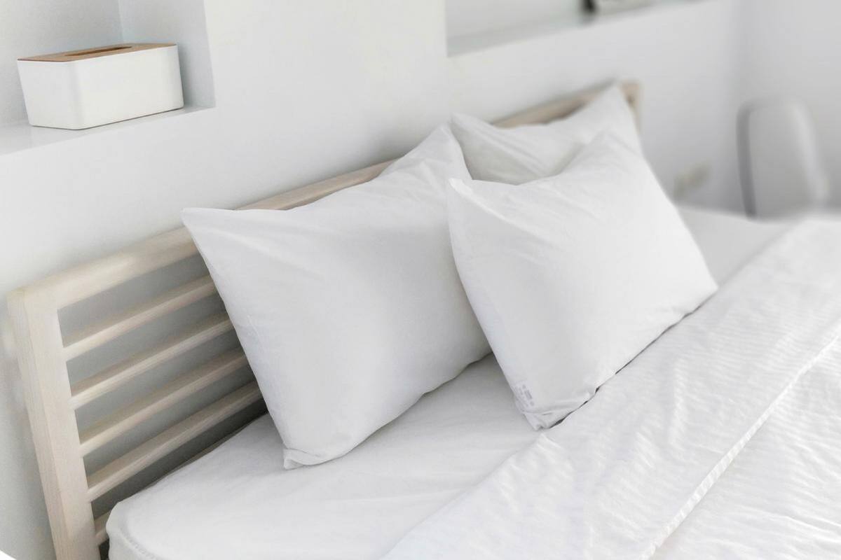A neatly made bed with white bedding and pillows, a wooden headboard, and a white bedside table with a small object on it.