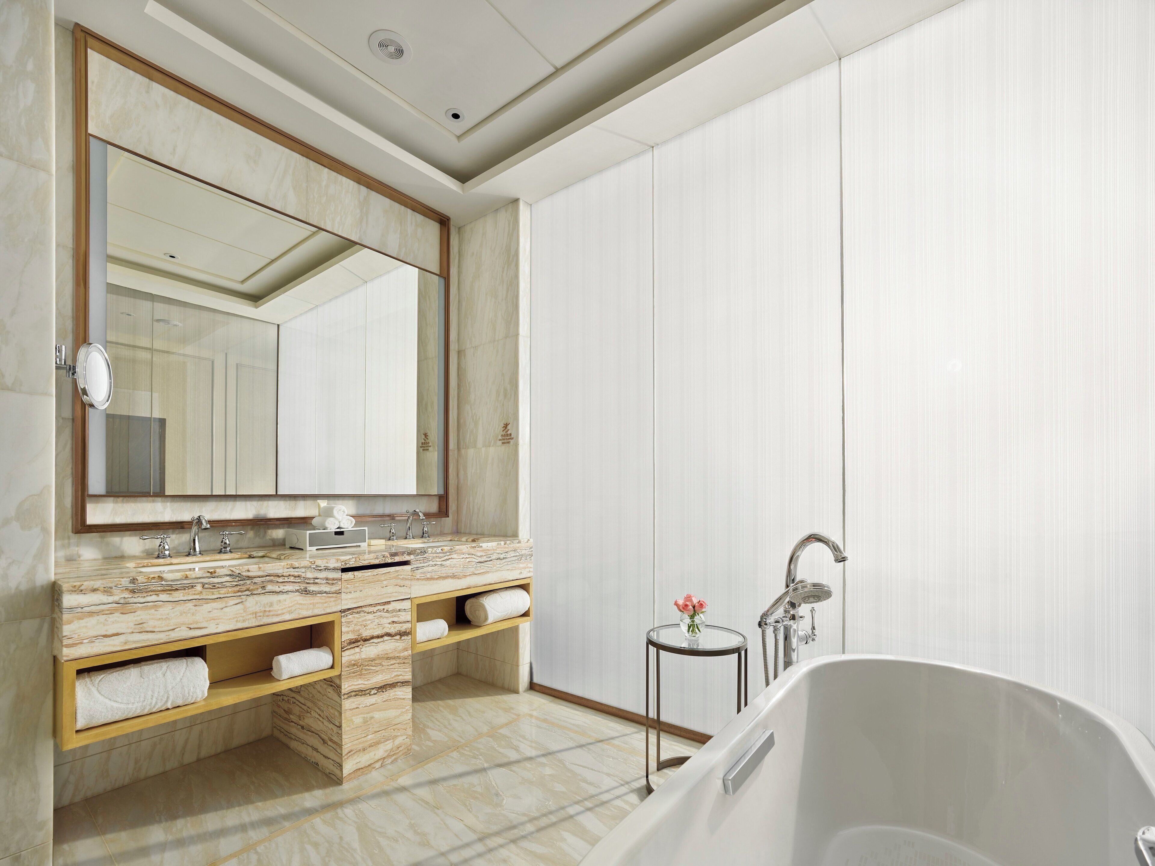 Een moderne badkamer met een marmeren wastafelmeubel, een grote spiegel, een vrijstaand bad en een klein tafeltje met een vaas.