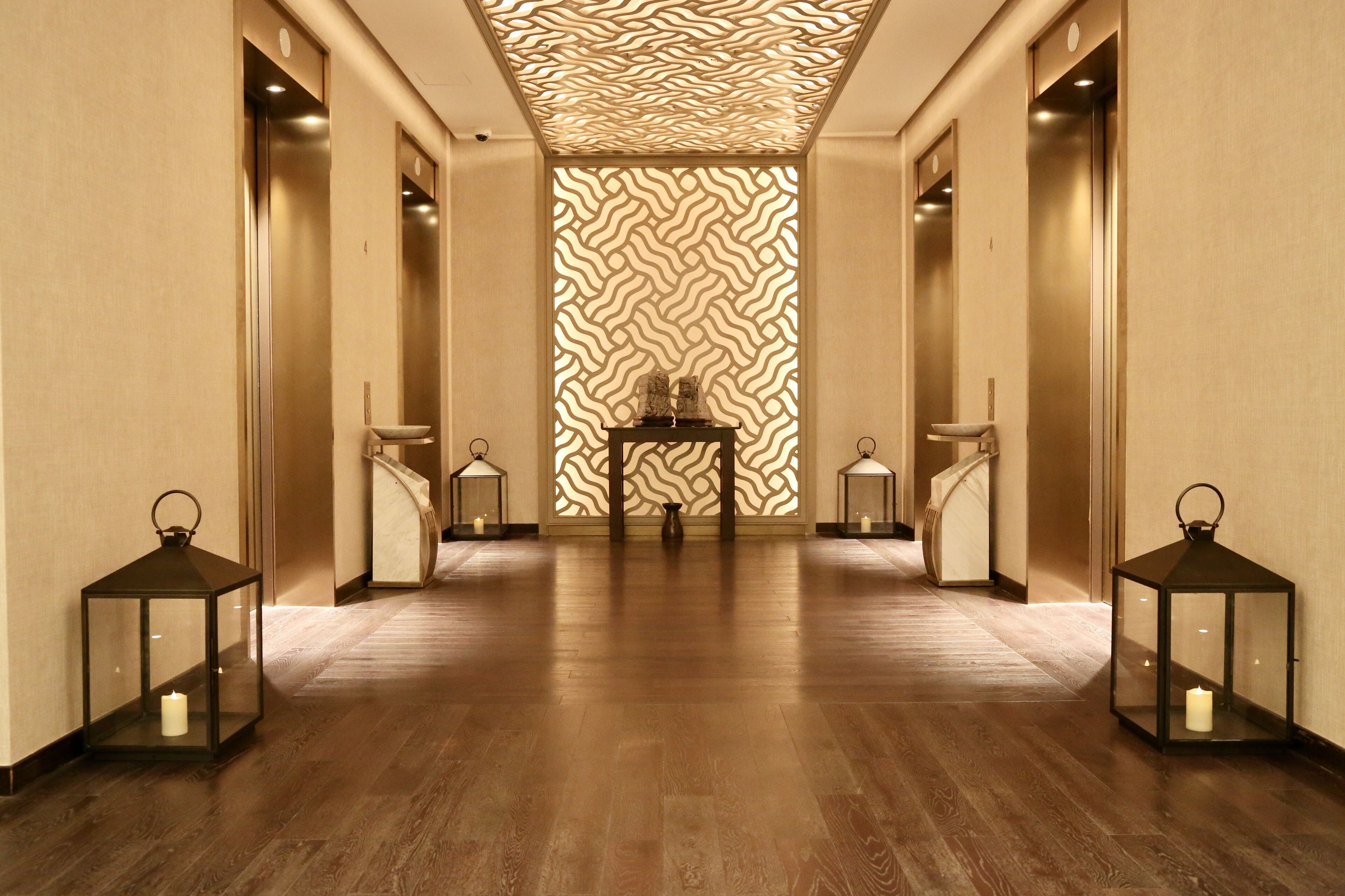 A hallway with wooden flooring, two side-by-side elevators, and a patterned wall in the background.