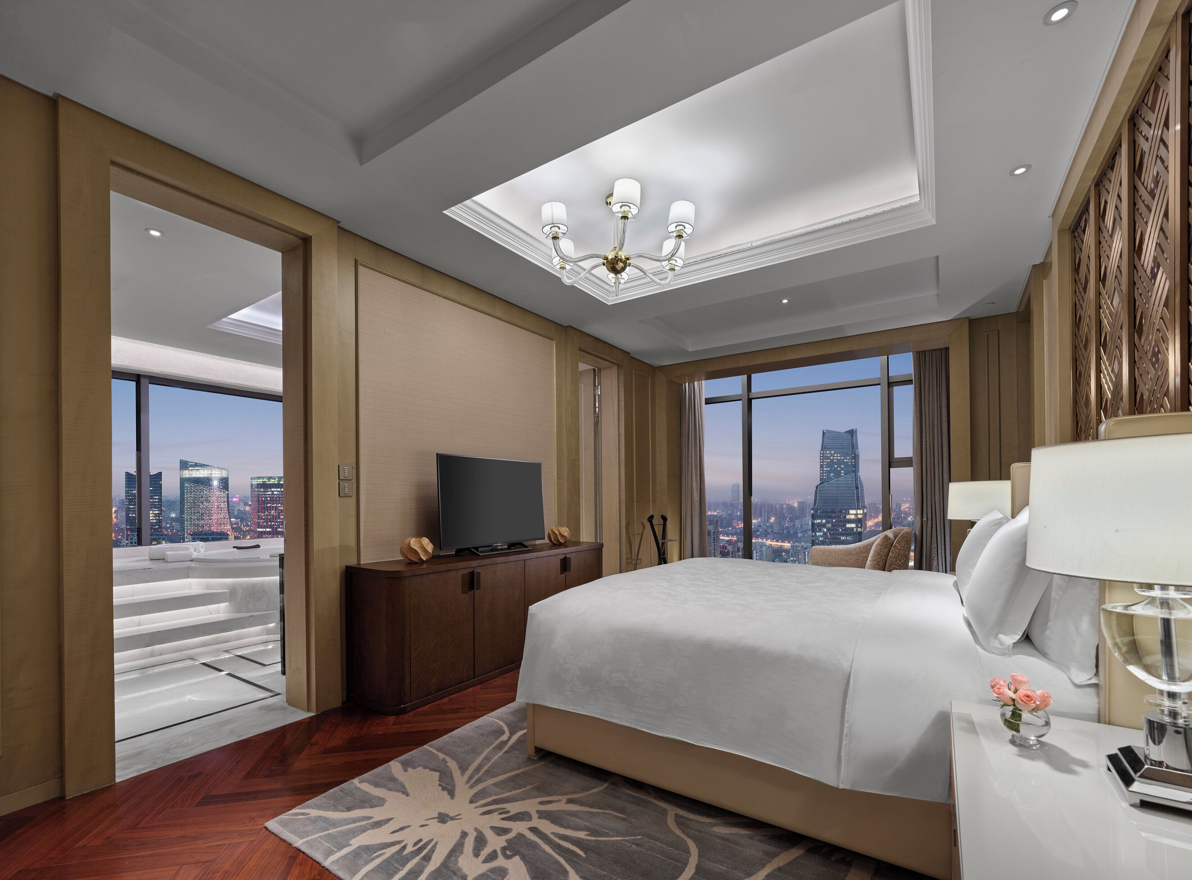 A spacious hotel room with a large bed, a flat-screen TV, a desk with a lamp, and a view of the cityscape through large windows.