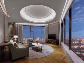 A modern hotel room with a large window offering a city view, a round ceiling light, a flat-screen TV, and a seating area with a coffee table.