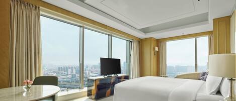 Habitación de hotel con una cama grande, un escritorio con televisor, una mesa de comedor y vistas a la ciudad.