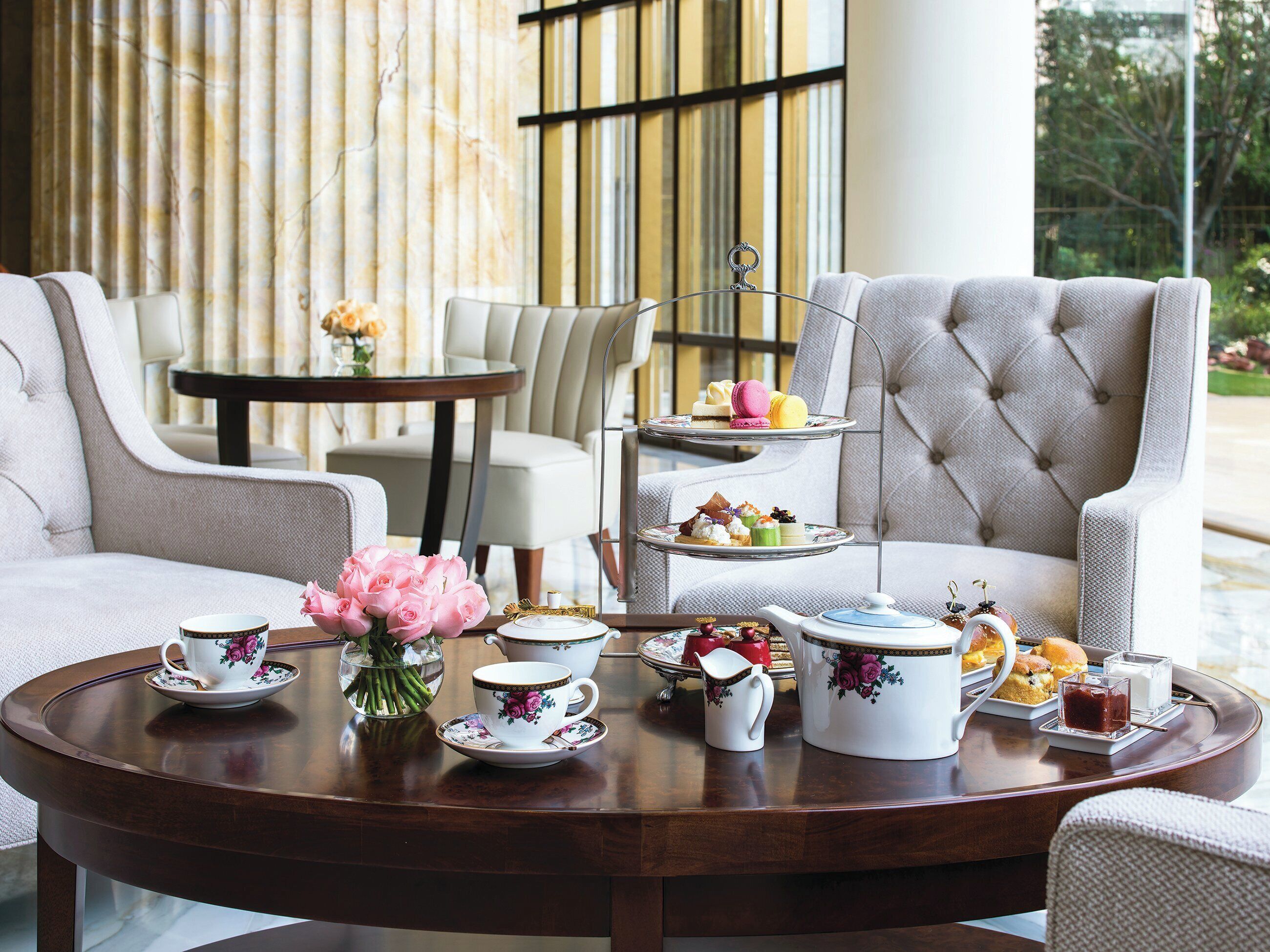 A three-tiered tea service set on a round table in a well-appointed room with a large window and a view of greenery outside.