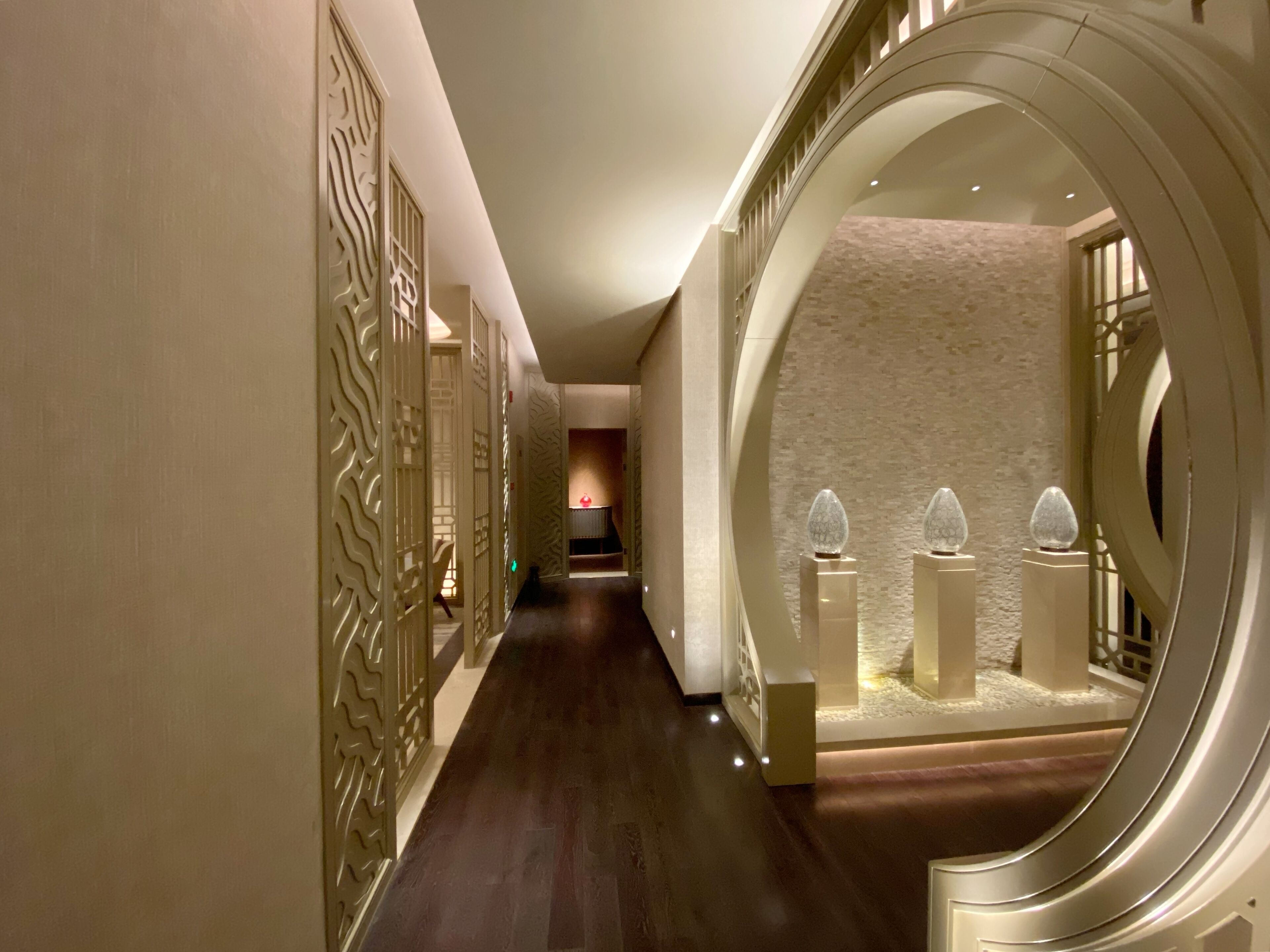 A corridor with ornate wall panels, a polished wooden floor, and a circular mirror with decorative elements.