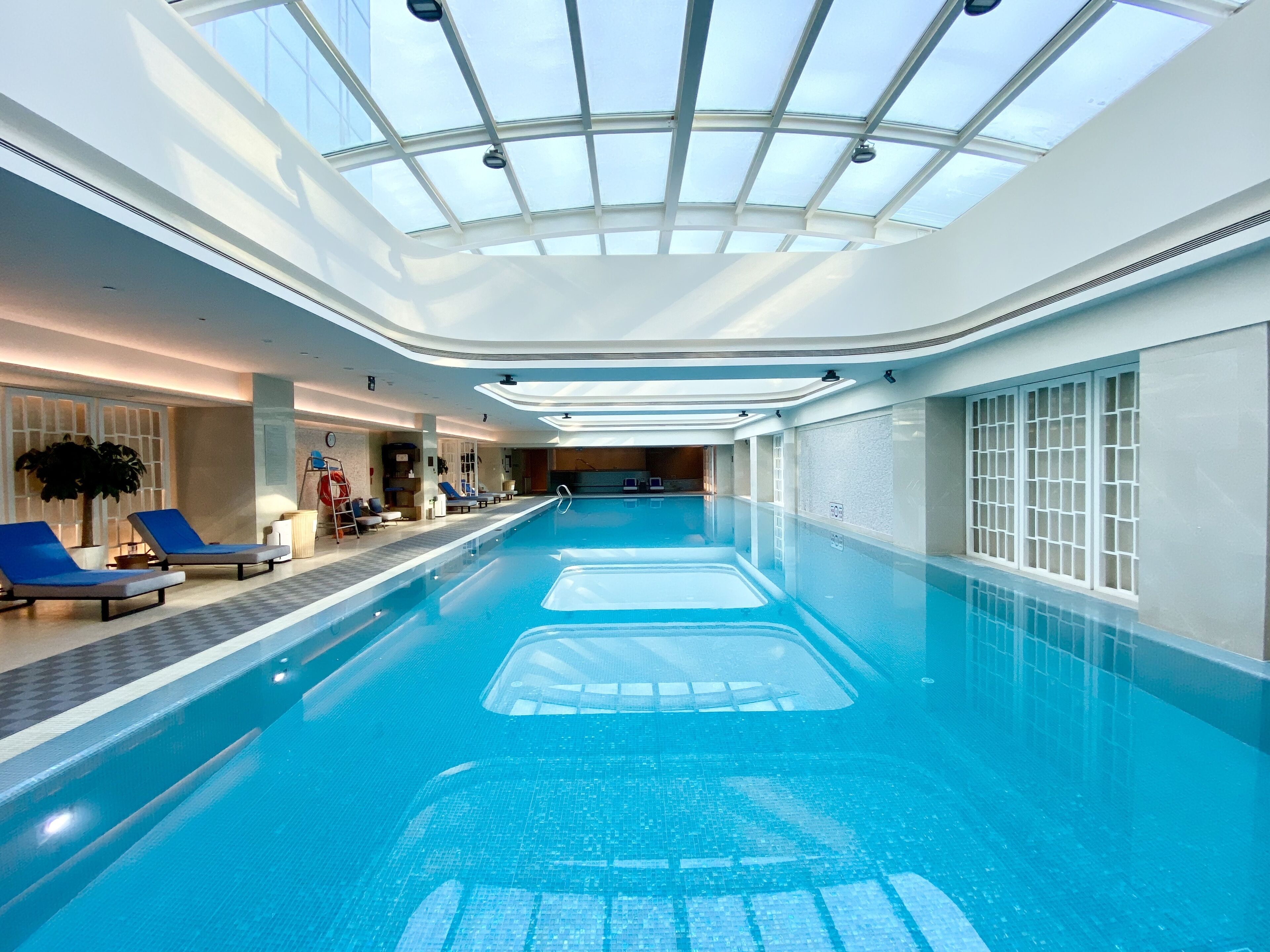 Une piscine intérieure dotée d’un plafond en verre transparent, de chaises longues bleues et d’un sol carrelé.