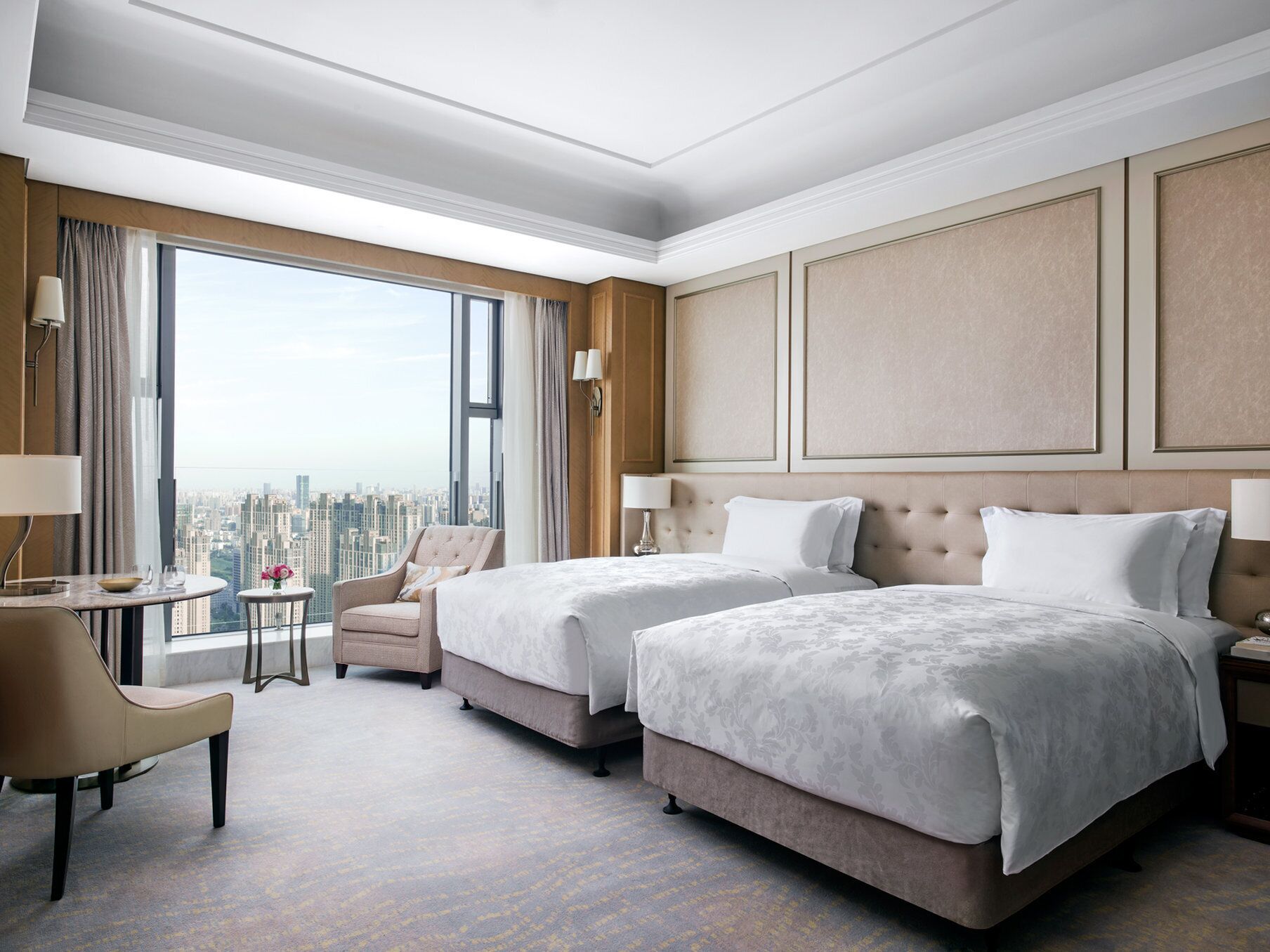 Habitación de hotel con dos camas, un área de descanso con una silla y una mesita, y un amplio ventanal con vistas a la ciudad.