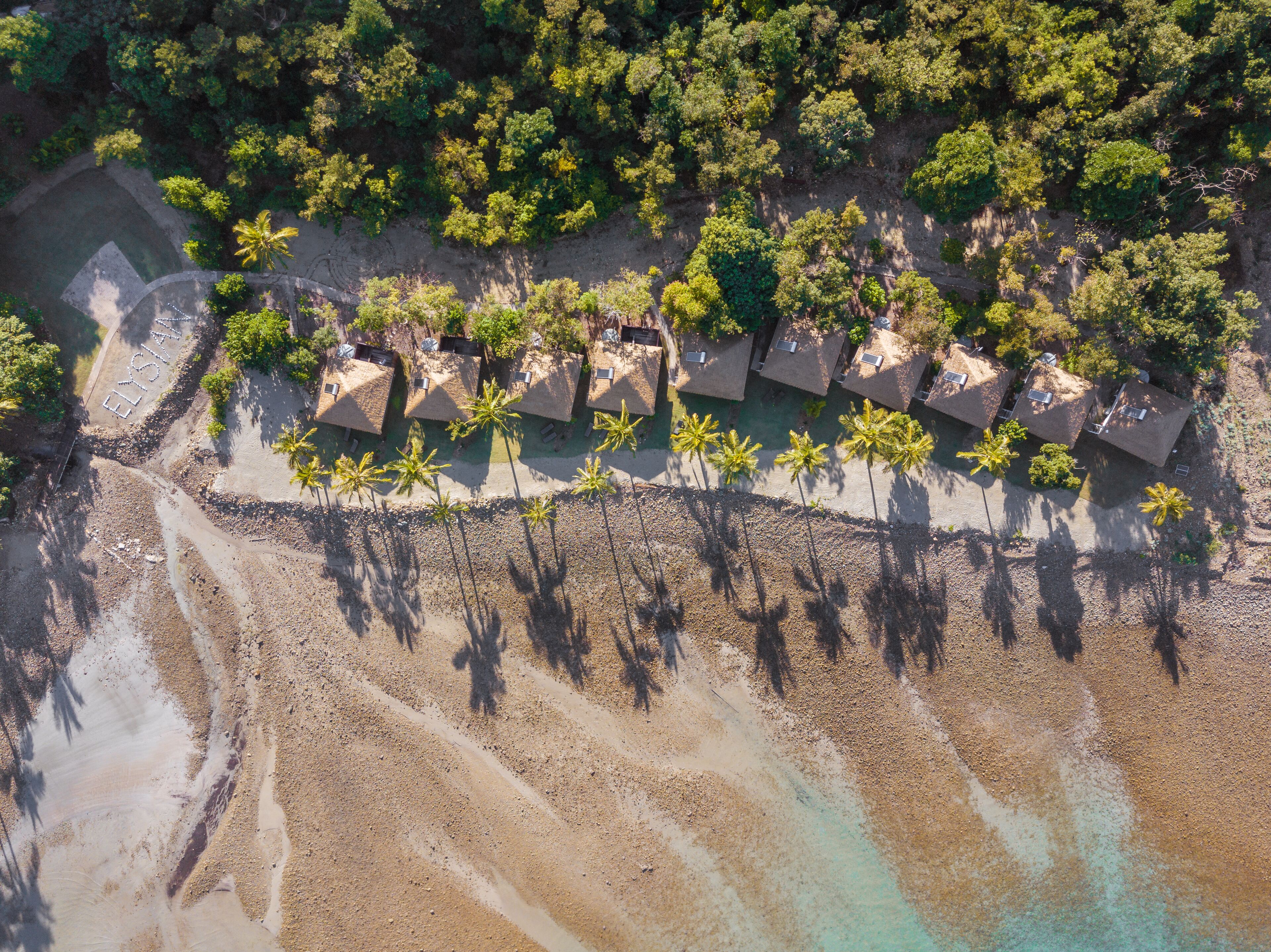 Luchtbeeld van een resort met rieten bungalows langs een strand, omgeven door weelderige tropische begroeiing.