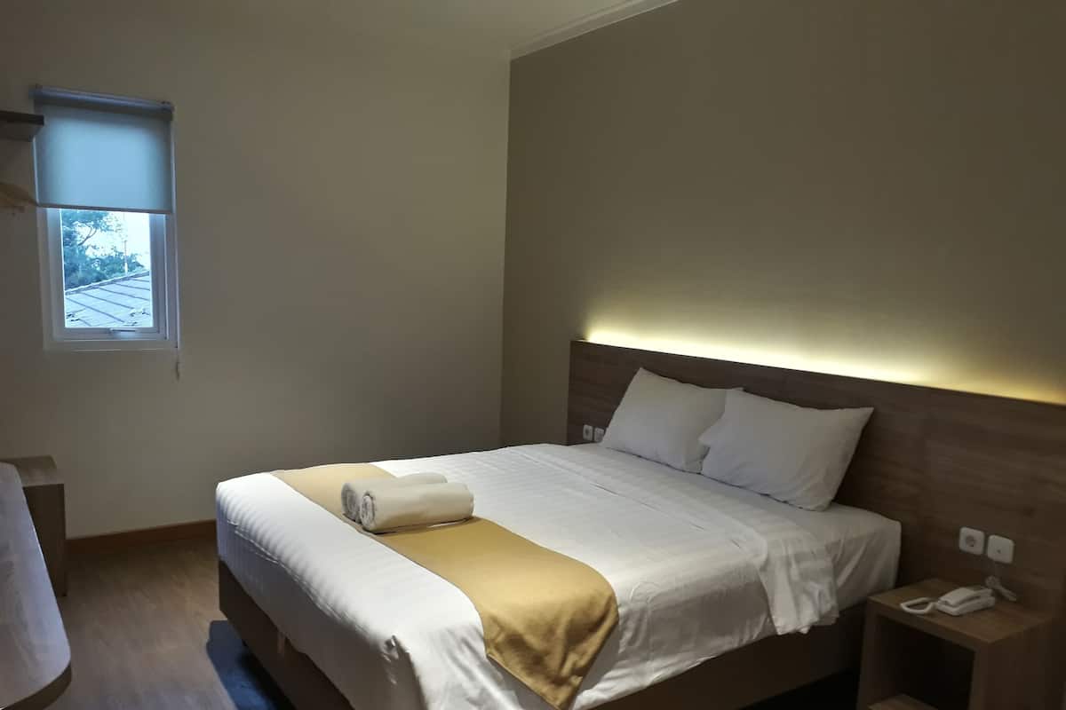 A neatly made bed with white and beige bedding, a bedside table with a phone, and a window with blinds.