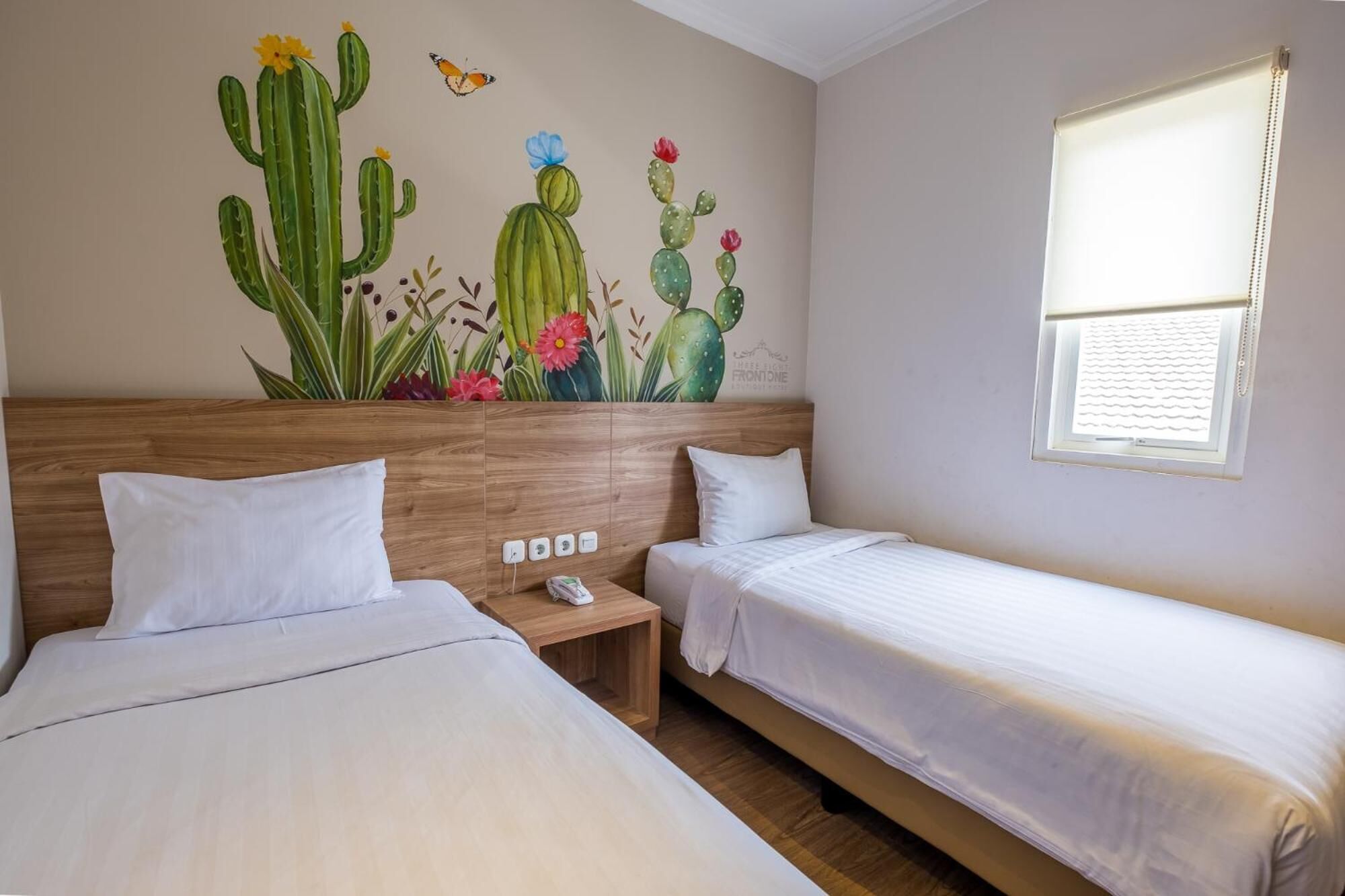 Een hotelkamer met twee bedden, een houten hoofdbord, een muurschildering van cactussen en bloemen, een raam met luiken en een nachtkastje met een telefoon.