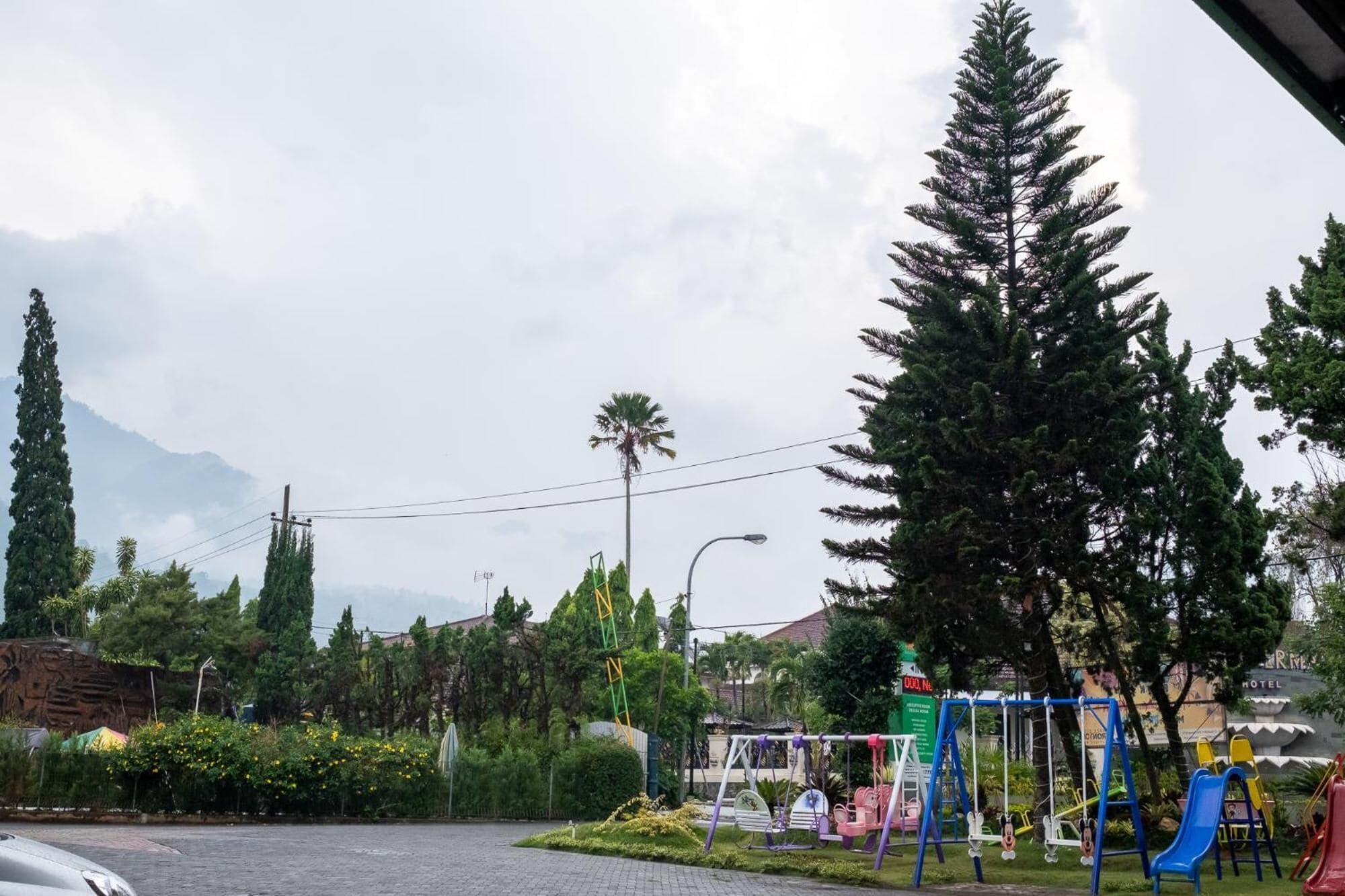 Un parque infantil con columpios y toboganes, rodeado de árboles y un hotel al fondo.