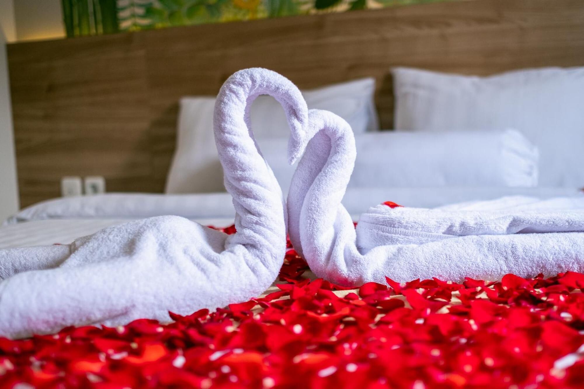 A hotel room with a bed covered in white towels arranged in a heart shape, surrounded by red rose petals.