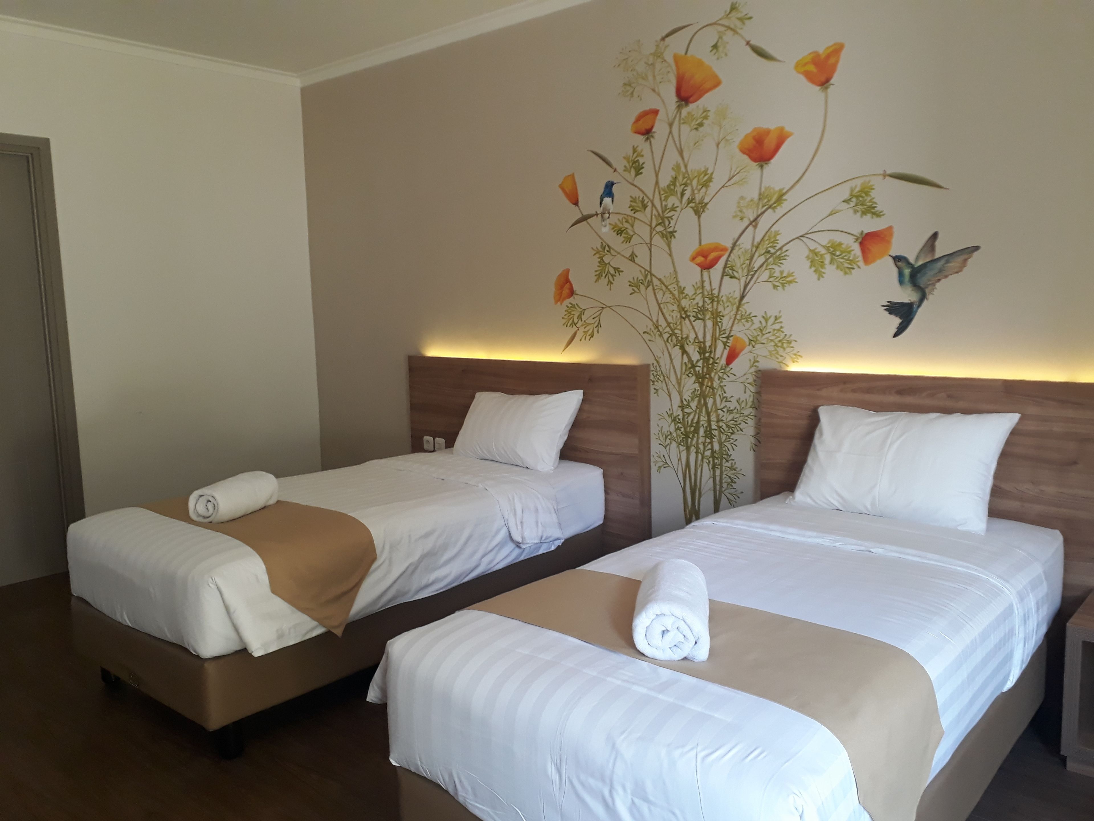 A hotel room with two single beds, each with white linens and a brown throw blanket. The room features a wooden headboard and a wall mural of birds and flowers.