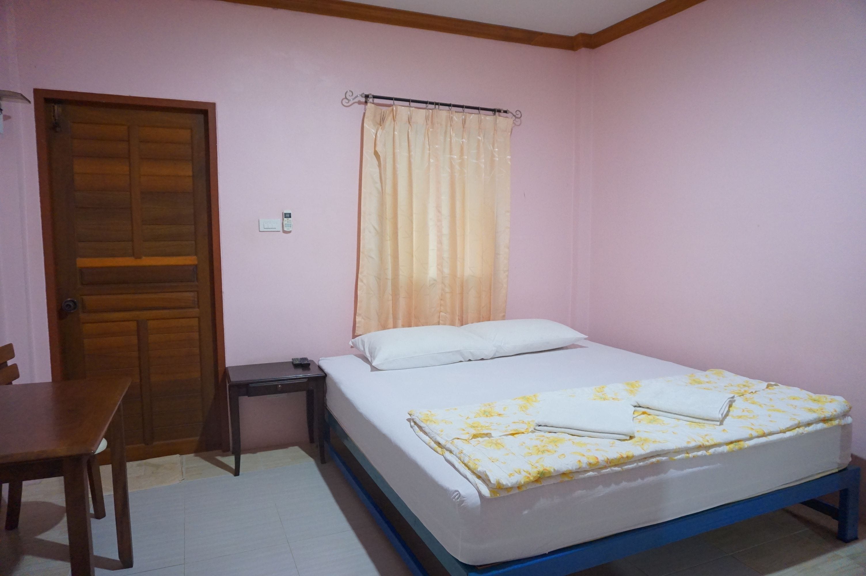 A bedroom with a wooden door, a bedside table, a bed with a floral bedspread, and a window with a curtain.