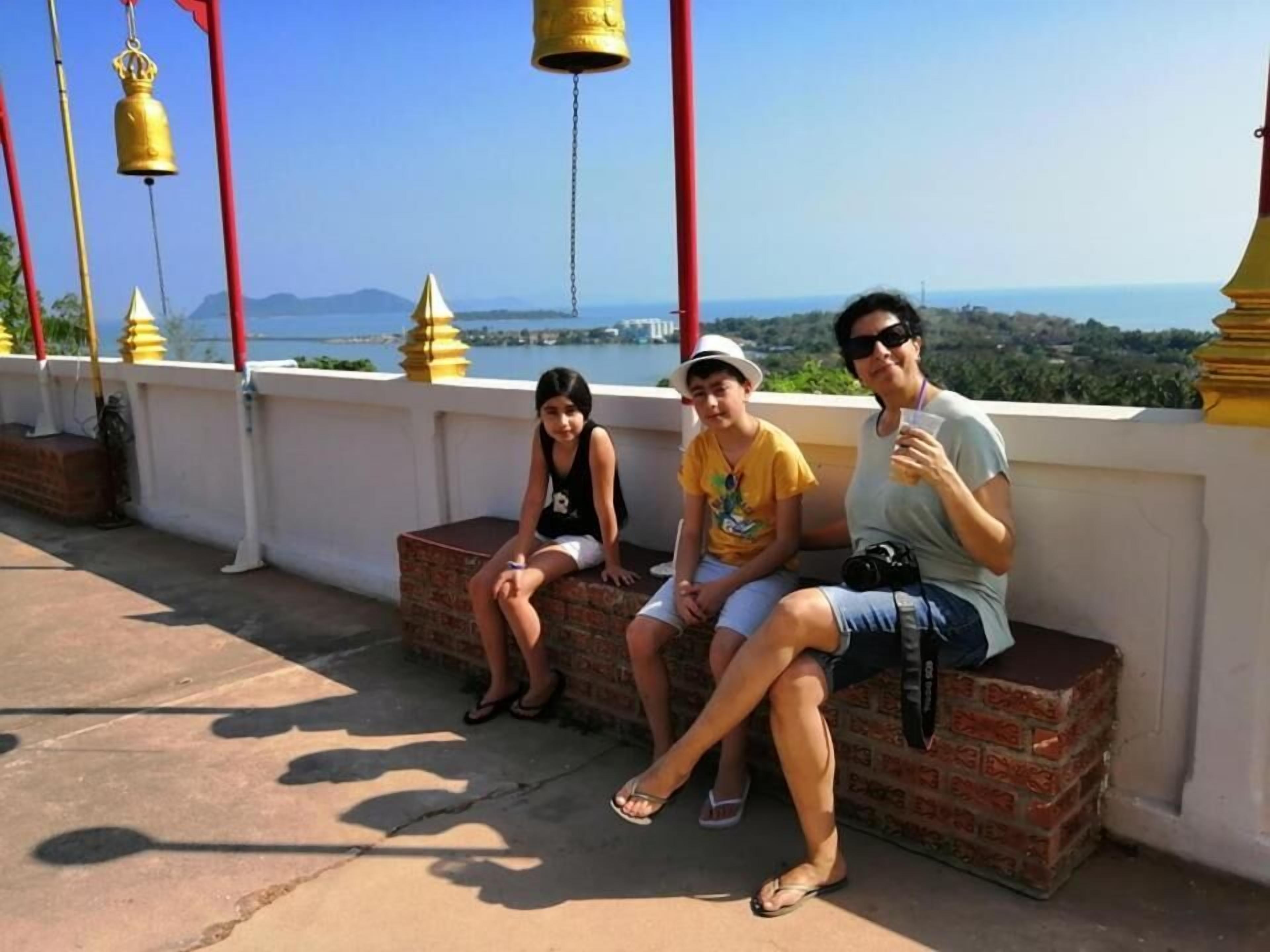 Drei Personen sitzen auf einer Bank mit Blick auf das Meer und die dahinterliegenden Berge.