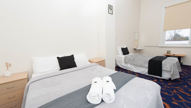A hotel room with two beds, a bedside table, a nightstand, a window with a blind, and a wall-mounted air conditioning unit. - Riverina Hotel (Holbrook)