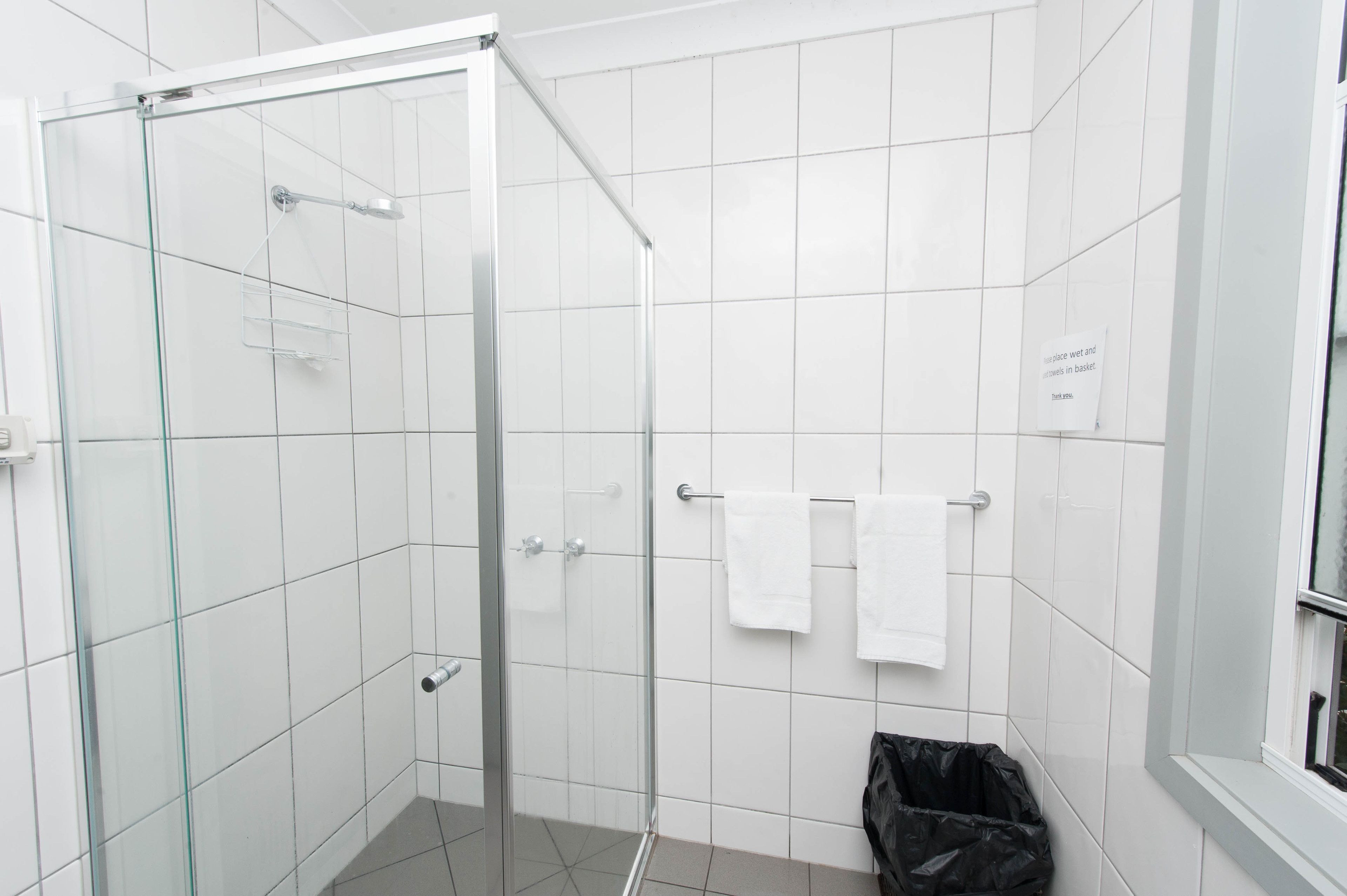 A modern bathroom with white tiles, a glass shower enclosure, and a black trash bin.