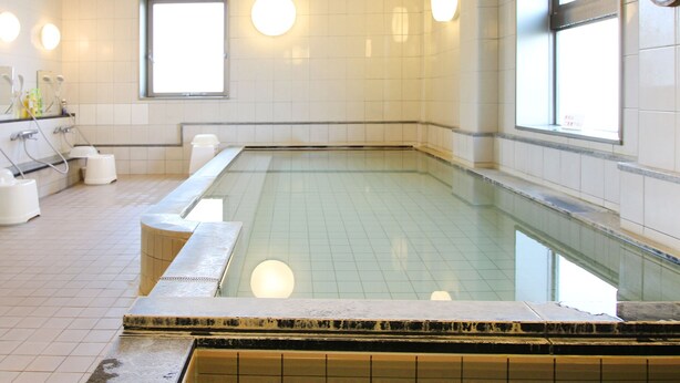 Un área de piscina cubierta limpia y bien iluminada, con paredes de azulejos, un largo banco a lo largo de la piscina y una fila de lavamanos con dispensadores de jabón.