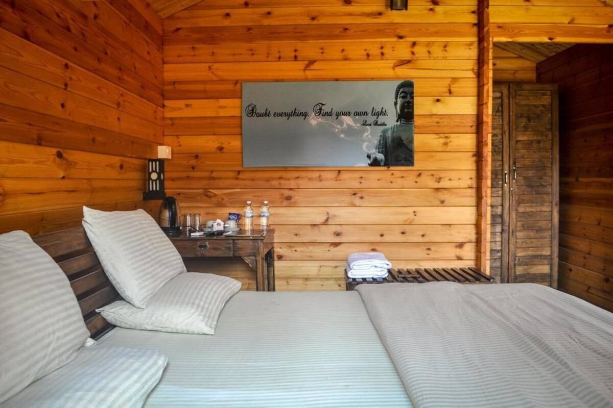 A cozy wooden cabin room with two beds, a small table with toiletries, and a motivational quote on the wall.