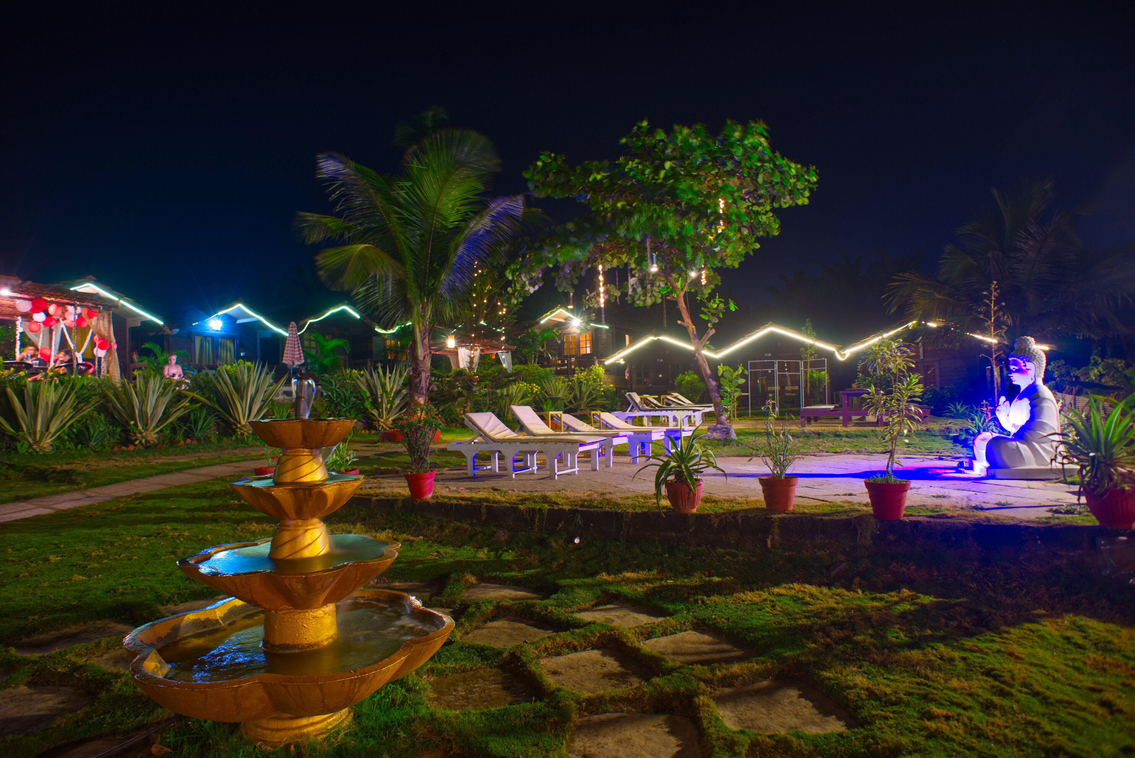 A nighttime resort with a fountain, lounge chairs, and potted plants.