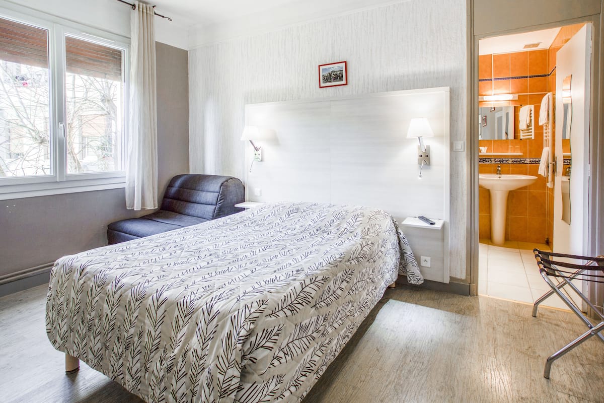A bedroom with a bed, a chair, a nightstand, a lamp, and a bathroom visible through an open door.