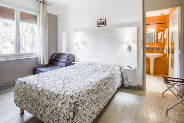 A bedroom with a bed, a chair, a nightstand, a lamp, and a bathroom visible through an open door. - Hôtel Le Provencal (Tarascon)