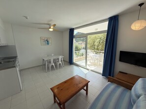A room with a dining table, chairs, a wooden bench, a sofa, a TV, a ceiling fan, and a balcony with a view.