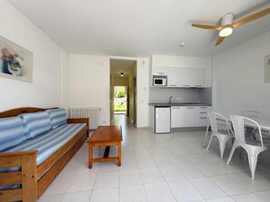 Un espace de vie compact comprenant un canapé, une table en bois, un coin repas avec des chaises, une kitchenette et un ventilateur de plafond.