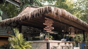 Un área de bar al aire libre rústica con techo de paja, mostrador de ladrillo y una pizarra que anuncia diversas bebidas.