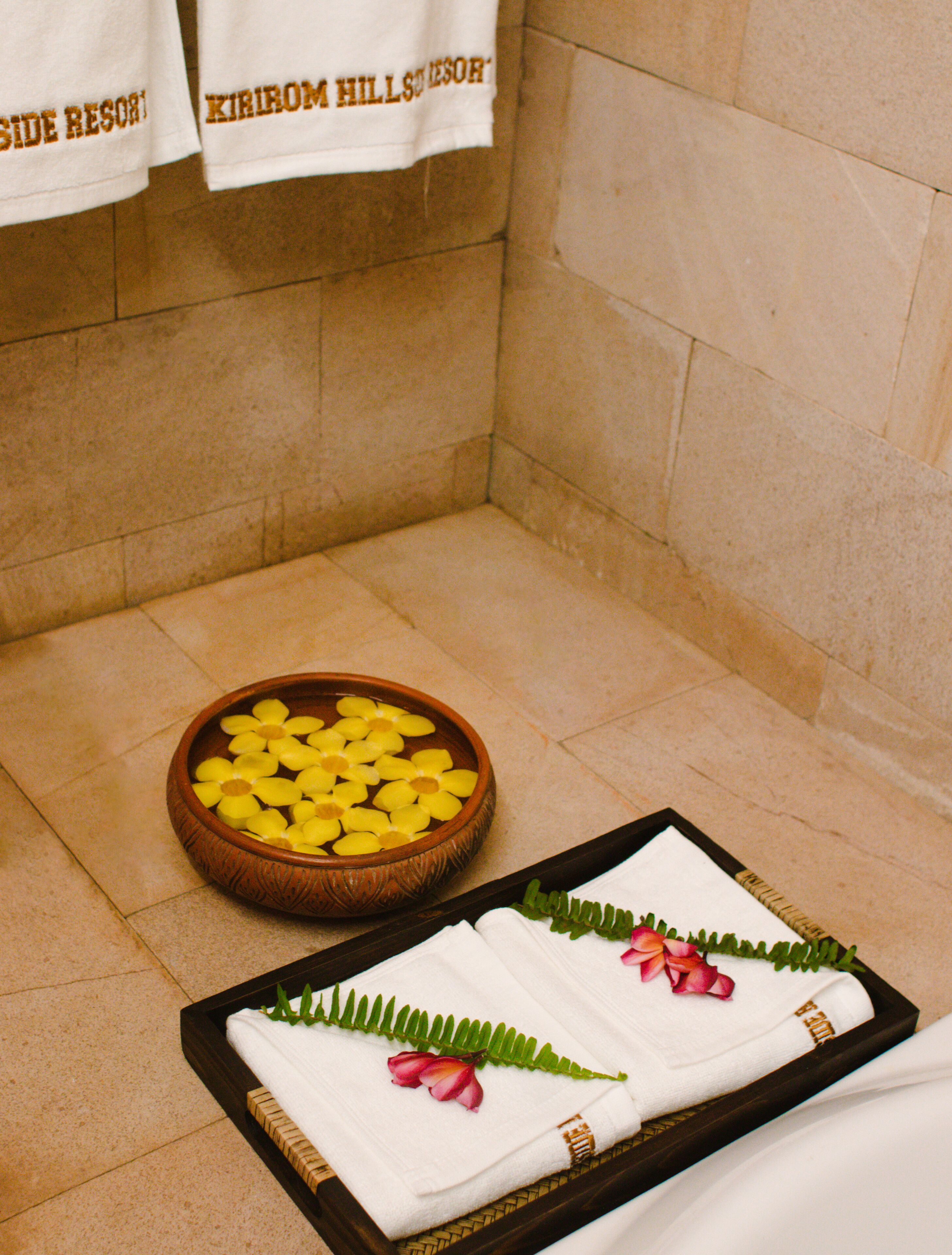 A bathroom with a wooden bowl of yellow flowers, two white towels with pink flowers, and towels hanging on the wall.