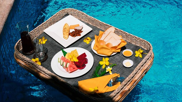 Una mesa de comedor junto a una piscina con una bandeja de mimbre que contiene comida, bebidas y cubiertos.