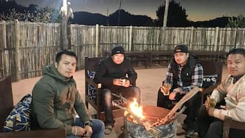 Drei Personen sitzen an einem Lagerfeuer im Freien, im Hintergrund ist ein Holzzaun zu sehen.