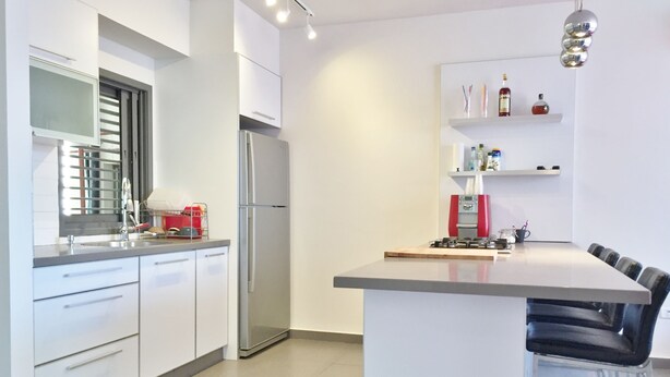 A modern kitchen with white cabinets, a stainless steel refrigerator, a coffee machine, and a bar area with stools. - Hilton Beach Stunning Designer 2BR APT (Tel Aviv)